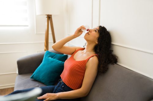 Tired young woman resting on the sofa putting her head back with a nose bleeding and using a tissue for her nosebleed