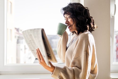 Beautiful young woman drinking cup of coffee reading newspaper