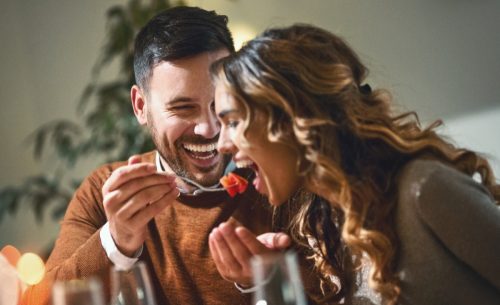  of mid 20's couple having fun during dinner party. The guy is feeding his girls with some chopped fruit, both laughing.