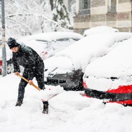 A man shoveling snow during a storm near a lineup of cars.