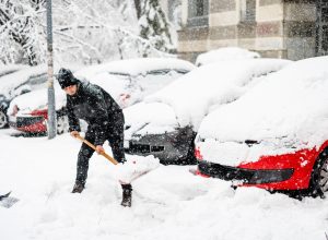A man shoveling snow during a storm near a lineup of cars.