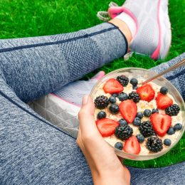 View of woman's legs in gray workout leggings while sitting in the grass holding a bowl of oatmeal and berries