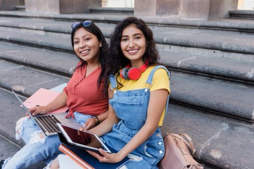 two young women sitting on university steps with laptop and tablet