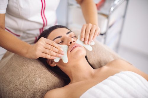 Beautiful mid adult woman receiving face massage with jade gua sha stones at salon