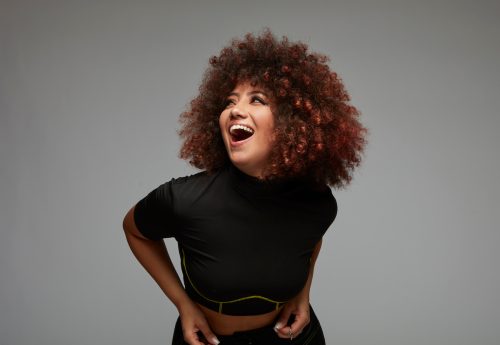 Laughing curly-haired young woman wearing black clothes against gray background