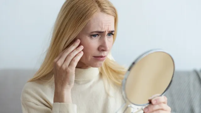 An upset middle-aged blonde woman wearing a pale yellow sweater looks at her face in a handheld mirror