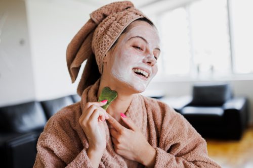 A young happy woman wearing a robe and hair towel has on a face mask and uses a gua sha tool on her neck