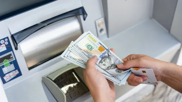 Closeup of hands holding 100 dollar bills and a debit card at an ATM