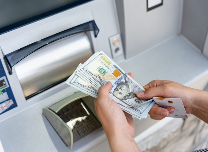 Closeup of hands holding 100 dollar bills and a debit card at an ATM