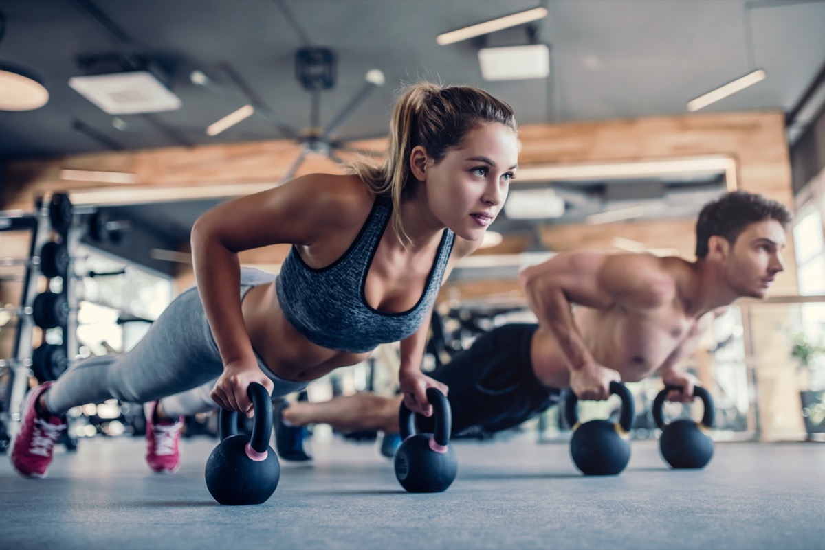 Young couple is working out at gym. Attractive woman and handsome muscular man are training in light modern gym. Doing plank on kettlebell. Push-up on weights.