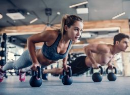 Young couple is working out at gym. Attractive woman and handsome muscular man are training in light modern gym. Doing plank on kettlebell. Push-up on weights.