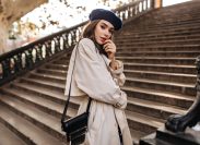 Lovely young Parisienne with brunette hair in stylish beret, beige trench coat and black bag, standing on old stairs and sensitively posing outdoors.