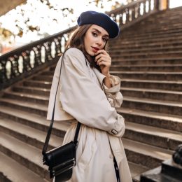 Lovely young Parisienne with brunette hair in stylish beret, beige trench coat and black bag, standing on old stairs and sensitively posing outdoors.