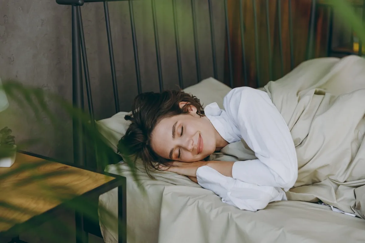 Young calm-looking woman sleeping in bed in sage-green sheets wearing a white sleep shirt