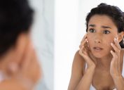 Atopic Skin Concept. Worried Young Woman Looking At Mirror And Touching Face, Unhappy Millennial Lady Suffering Dermatitis, Examining Irritation Signs While Standing In Bathroom At Home, Closeup