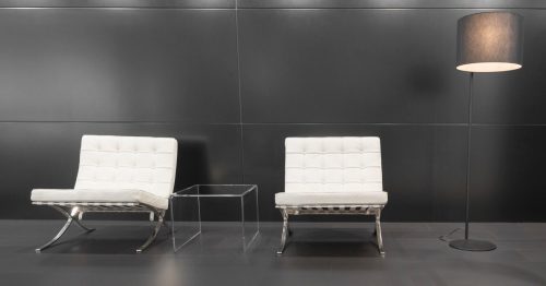 Pair of white modernist Barcelona design chairs and a modern lamp in front of a grey background. The Barcelona chair was deisgned by Ludwig Mies van der Rohe and Lilly Reich for the German pavillion for the International Exposition of 1929 in Barcelona.
