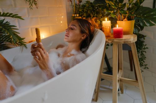 Photo of a young woman using a phone while taking a bubble bath; checking her social media and news online, relaxing, and having a spa day at home.
