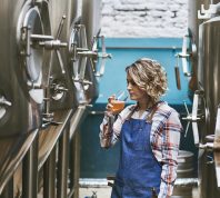 A female brewer sampling her beer while standing next to fermenters in a brewery