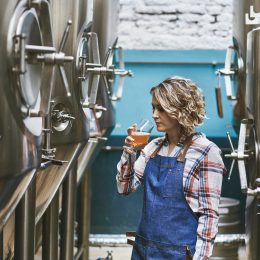 A female brewer sampling her beer while standing next to fermenters in a brewery