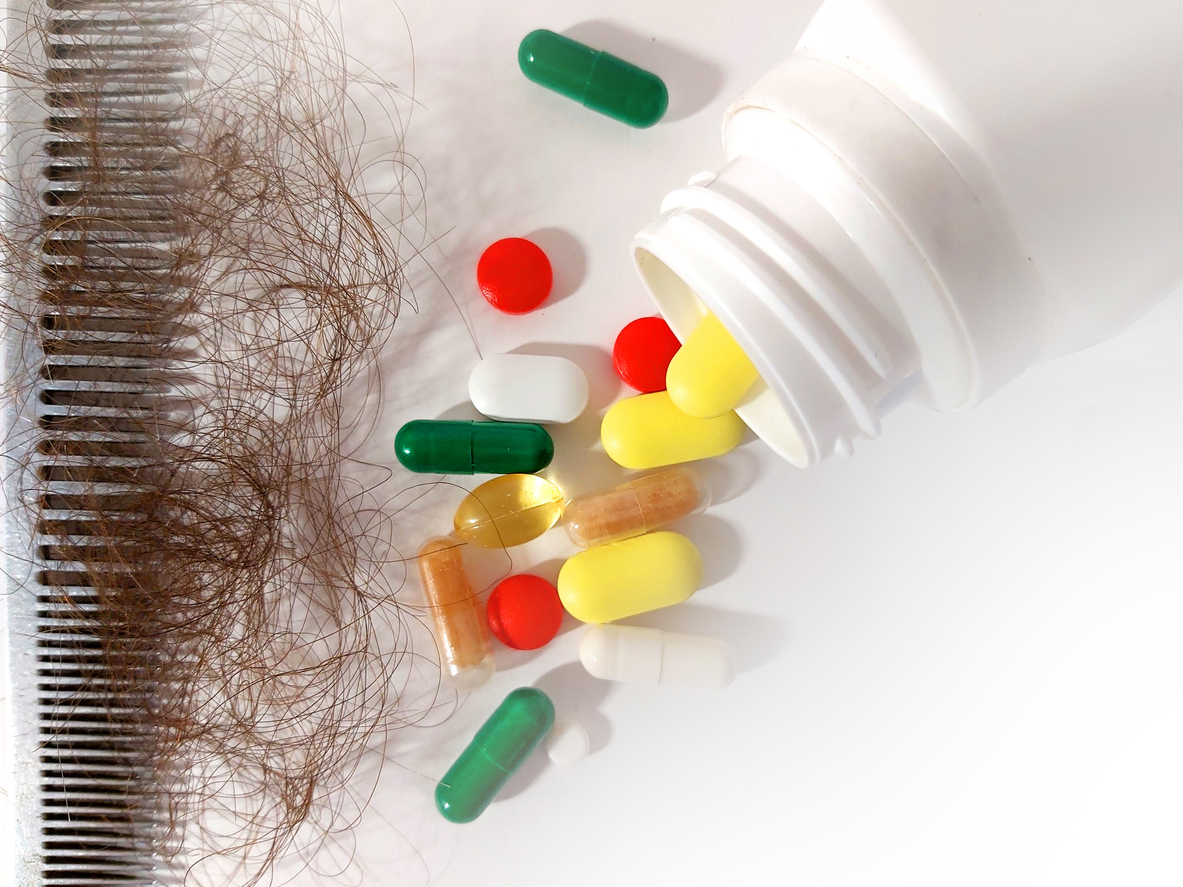 A comb full of brown hair next to a spilled out bottle of supplements on a white background