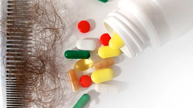 A comb full of brown hair next to a spilled out bottle of supplements on a white background