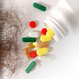 A comb full of brown hair next to a spilled out bottle of supplements on a white background