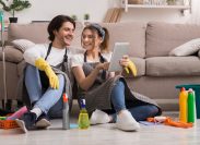 Cheerful Young Couple Relaxing With Digital Tablet while sitting on floor in front of couch surrounded by cleaning supplies