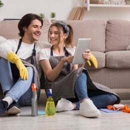 Cheerful Young Couple Relaxing With Digital Tablet while sitting on floor in front of couch surrounded by cleaning supplies