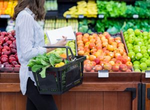 Young woman carries a shopping basket filled with fresh produce. She is shopping for fresh fruit and vegetables in a grocery store.