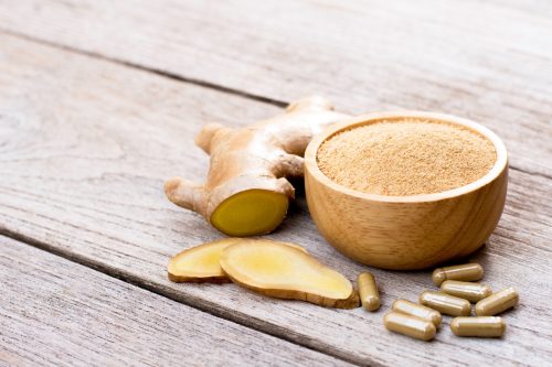 Fresh ginger slice and powder capsules with ginger ground in wooden bowl isolated on wood table background. Herbal medicine plant and supplement concept.