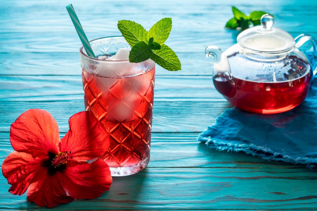 A hibiscus tea cocktail in a glass next to a clear kettle