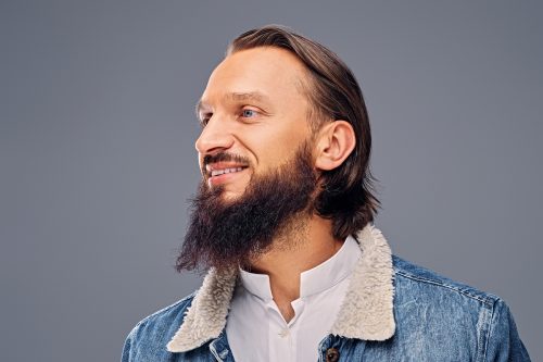 Portrait of bearded male with long hair dressed in a denim jacket over grey background.