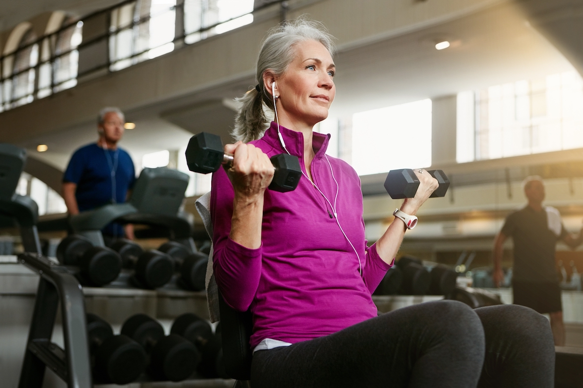 Mature woman with gray hair wearing a purple top using hand weights at the gym