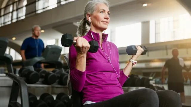 Mature woman with gray hair wearing a purple top using hand weights at the gym