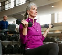 Mature woman with gray hair wearing a purple top using hand weights at the gym