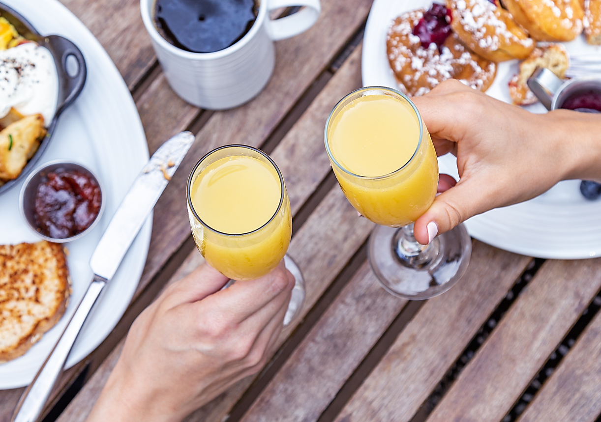 An overhead shot of two people cheersing with Mimosas at a brunch table