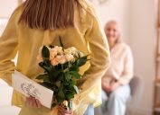 view from behind of young woman holding flowers and a mother's day card behind her back as she looks at an older woman