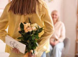 view from behind of young woman holding flowers and a mother's day card behind her back as she looks at an older woman