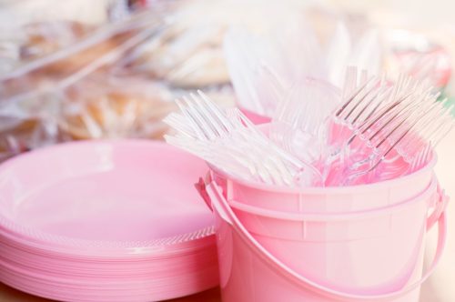 A stack of Pink Plates and a pink bucket filled with forks and knives sitting on a table. Copy Space available.
