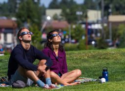 Man and woman sitting on grass while watching a solar eclipse with protective glasses