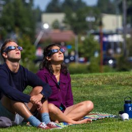 Man and woman sitting on grass while watching a solar eclipse with protective glasses