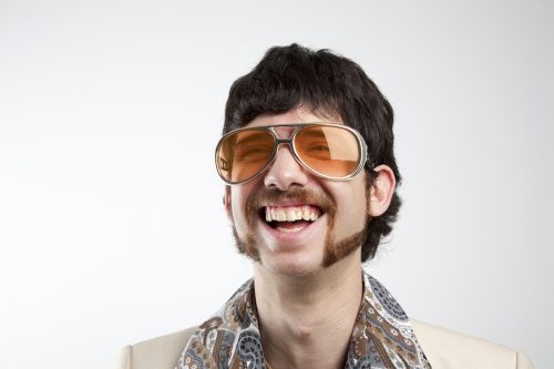 Close up portrait of a retro man in a 1970s leisure suit and sunglasses smiling and laughing with a mutton chop beard