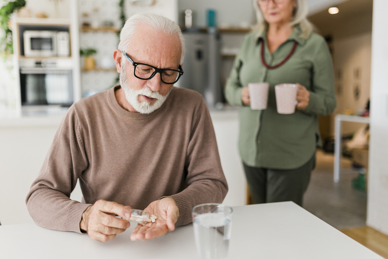 A senior man sitting at a table and taking vitamins or medicine while someone brings him a drink