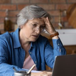 A senior woman sitting at her laptop in a kitchen with a distressed look on her face