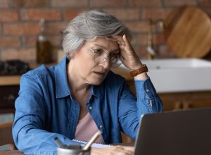 A senior woman sitting at her laptop in a kitchen with a distressed look on her face