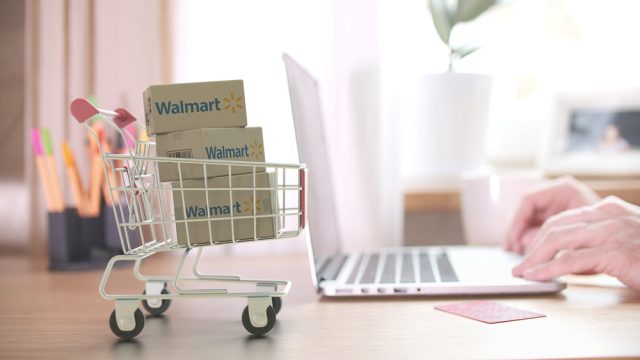 A miniature shopping cart filled with Walmart boxes next to someone using a laptop