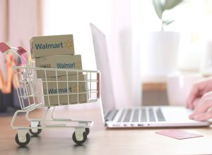 A miniature shopping cart filled with Walmart boxes next to someone using a laptop