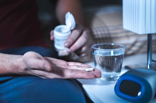A man sitting in bed and trying to take sleeping pills or night pills. He suffers from insomnia. He holds a tablet and a bottle in his hands. A glass of water on the nightstand in the bedroom.