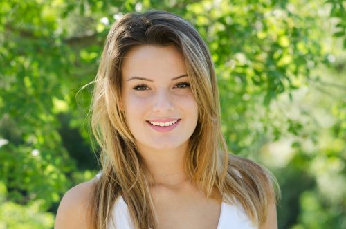 Young woman with long dirty-blonde hair smiling in front of a greenery background