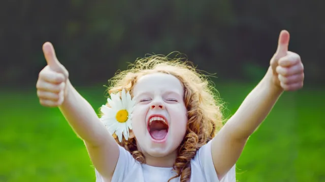 little girl with daisy in her hair making a double thumbs up and laughing at funny spring jokes